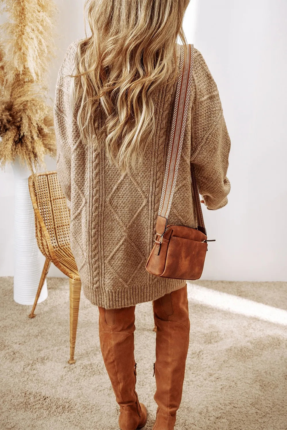 Person wearing a brown cable knit sweater and tan pants, holding a brown leather bag.