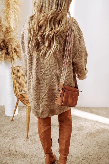 Person wearing a brown cable knit sweater and tan pants, holding a brown leather bag.